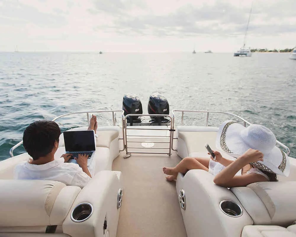 A man uses a laptop while a woman checks her phone on cushioned seats aboard a boat.